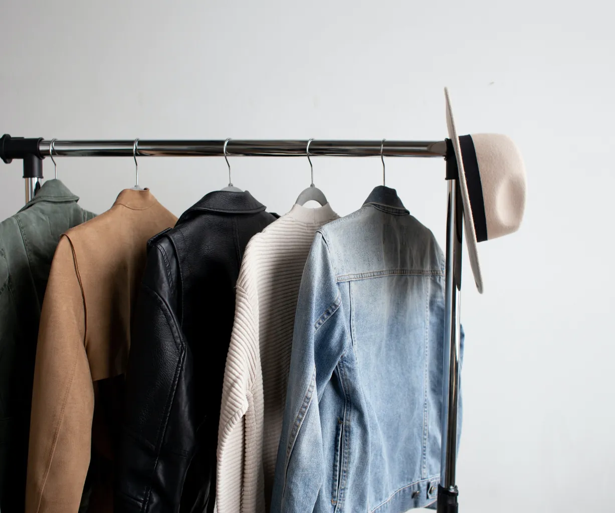 A clothing rack holds four jackets—tan, black leather, white knit, and blue denim—with a beige hat hanging on the rack’s end against a plain white wall.