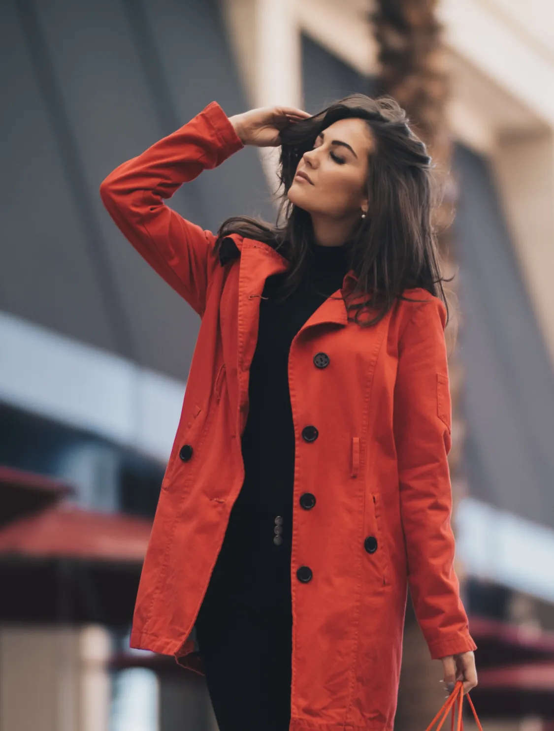 Woman wearing a red coat stands outdoors, eyes closed and hand in her hair, holding a small orange bag.