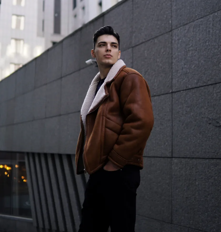 A person wearing a brown shearling jacket stands outdoors near a gray concrete wall, looking off to the side with a neutral expression.