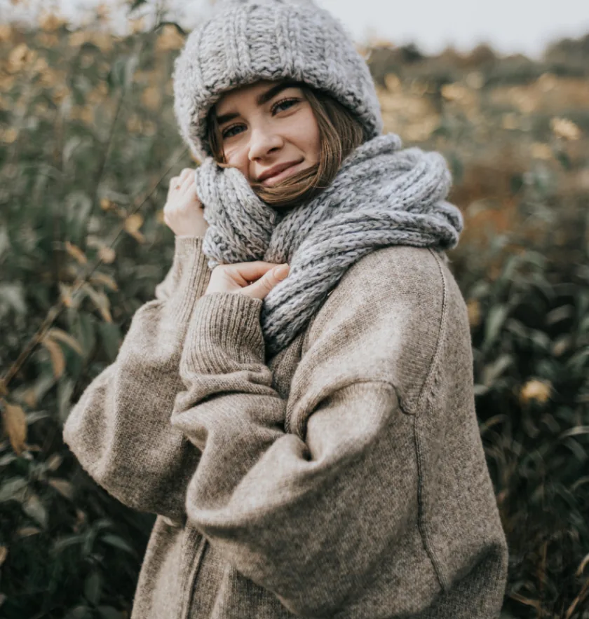 A person wearing a thick knit sweater, scarf, and hat stands outdoors in a grassy area, looking at the camera and smiling slightly.