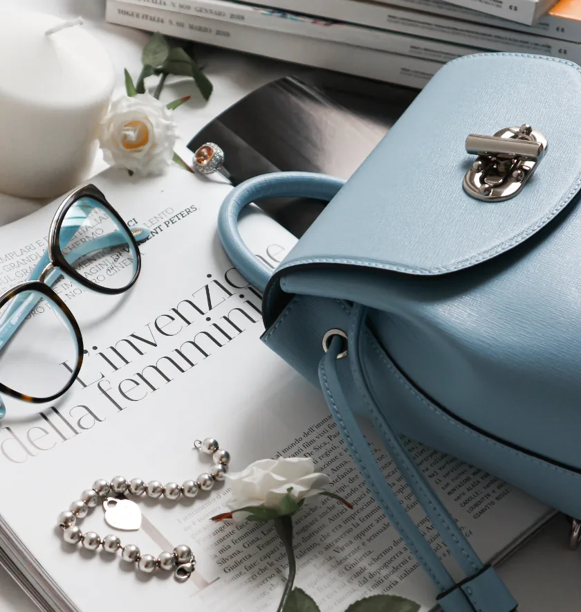 A light blue handbag, eyeglasses, a pearl bracelet, a white rose, and stacked magazines are arranged on a table.